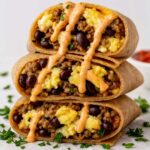 Stack of high protein breakfast burritos filled with scrambled eggs, black beans, ground meat, and sauce.