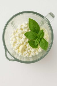 Glass blender bowl filled with cottage cheese, fresh basil leaves, a garlic clove, and grated Parmesan cheese, ready for blending.