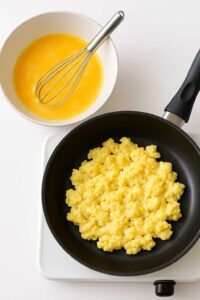 Bowl of whisked eggs with a whisk and scrambled eggs cooking in a nonstick skillet for high protein breakfast burrito