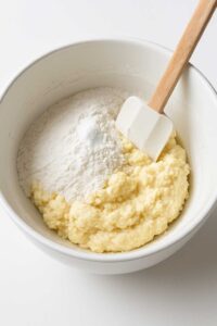 Flour and baking powder added to cottage cheese bagel mixture in a white bowl with a spatula