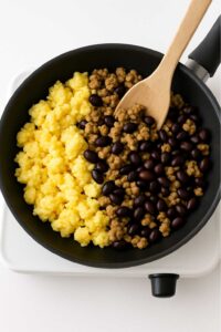 Scrambled eggs, turkey sausage, and black beans in a skillet with a wooden spoon high protein breakfast burrito meal prep​