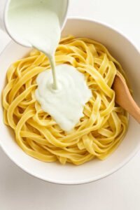Creamy cottage cheese pasta sauce being poured over cooked tagliatelle in a white bowl with a wooden spoon.