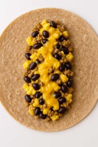 Whole wheat tortilla topped with scrambled eggs, turkey sausage, black beans, and melted cheese. for high protein breakfast burrito meal prep​