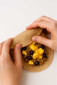 Hands rolling a whole wheat tortilla filled with scrambled eggs, turkey sausage, black beans, and melted cheese for the high protein breakfast burrito meal prep​