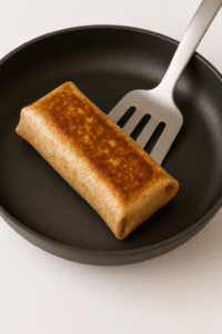 A golden-brown high protein breakfast burrito​ being toasted in a skillet with a spatula.