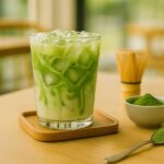 Tall glass of iced matcha latte with bright green swirls and ice cubes, set on a sunny café table with a bamboo whisk and spoonful of matcha powder nearby.