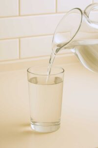Water being poured from a transparent pitcher into a clear glass on a neutral kitchen counter, step 1 of the pink salt diet recipe for weight loss.