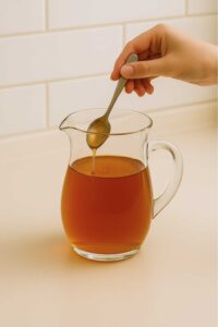 A hand stirs honey into a glass pitcher filled with warm amber apple peel recipe for weight loss, adjusting the sweetness for the weight loss recipe.