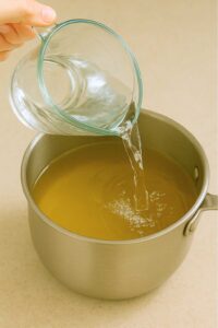 Hand pouring cold water from a glass measuring cup into a saucepan of green tea mixture for preparation green tea lemonade recipe